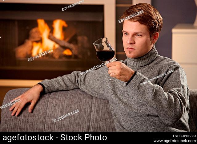 Young man sitting on sofa at home on a cold winter day in front of fireplace, drinking red wine