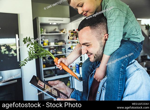 Smiling man using tablet while son sitting on shoulder in kitchen at home