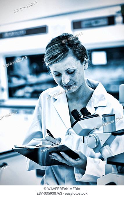 Serious female scientist writing on her clipboard in front of a microscope