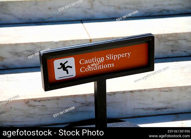 Caution, slippery conditions brown sign with icon representation on marble steps with a black holder on a sunny day