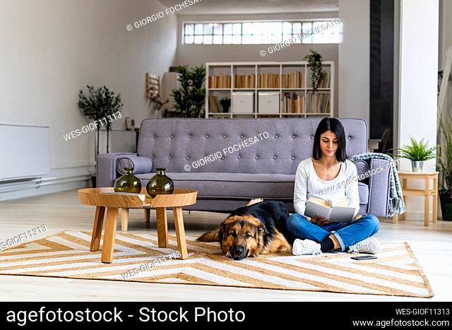 Woman reading book while sitting with dog against sofa at home