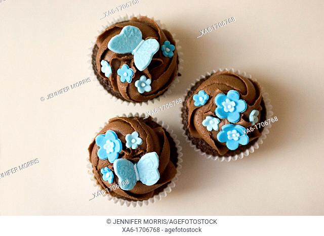 Three cupcakes with chocolate frosting swirl and blue butterfly and flower decorations on