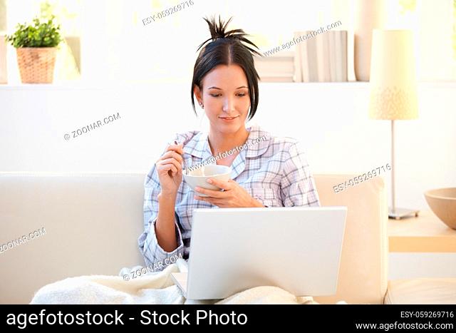 Girl in pyjama having cereal for breakfast, looking at laptop computer screen, smiling