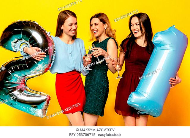 Young company of cheerful lovely girls in stylish dresses celebrating 21st anniversary, birthday party, holding helium balloons
