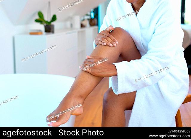 Mid section of woman in a bathrobe applying moisturizer on her leg sitting near bath tub