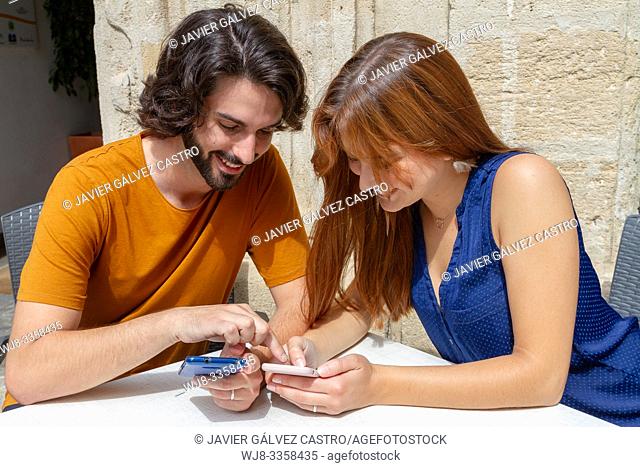 Young couple looking at the mobile on a summer terrace, smile while they wait