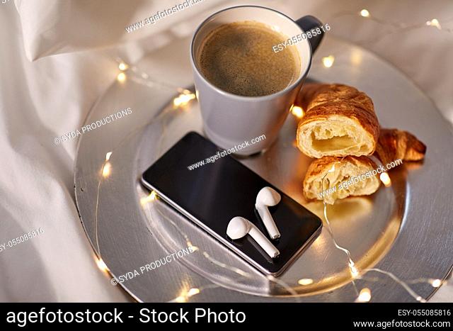 smartphone, earphones, coffee and croissant in bed