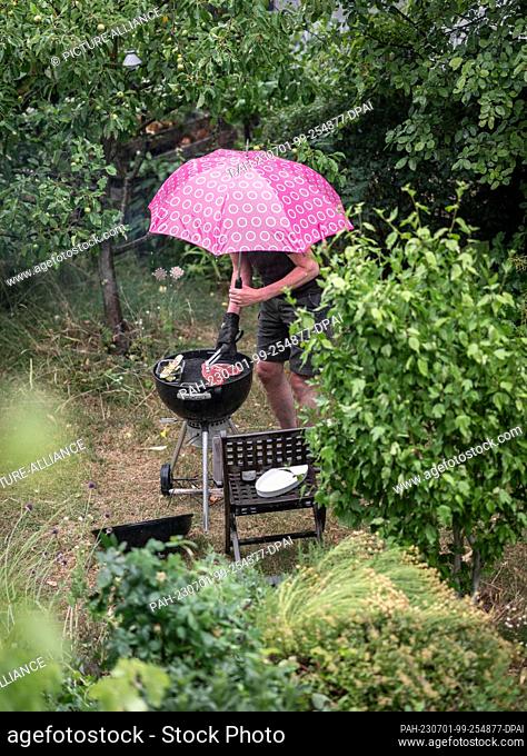 01 July 2023, Hesse, Frankfurt/Main: A man, caught in a rainstorm, stands in his backyard at a charcoal grill, turning meat on the grate