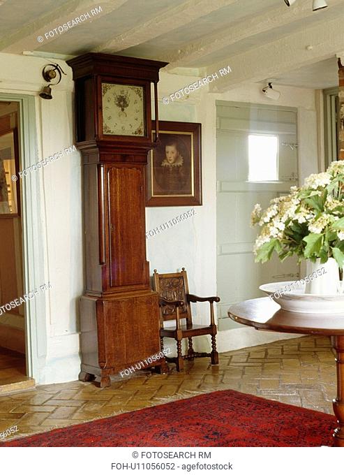 Antique longcase clock and oil portrait in cottage hall with white beamed ceiling and red rug on polished brick floor