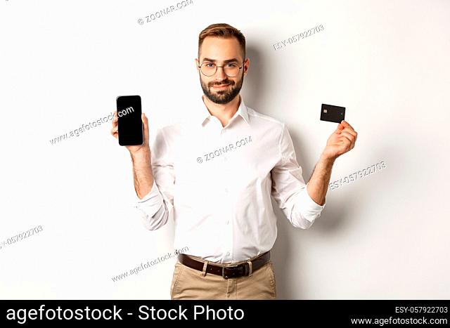 Handsome bearded man showing mobile phone and credit card, shopping online, standing over white background