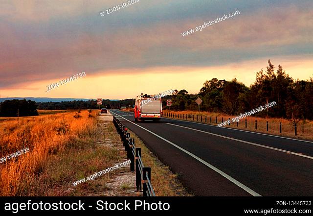 A Rural Fire Service, fire and rescue vehicle responds urgently to a large out of control bushfire in the mountains. Smoke and sun casts a red glow across the...