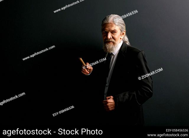 Elrderly stylish man in black jacket holding cigar. Old male portrait in studio on black background