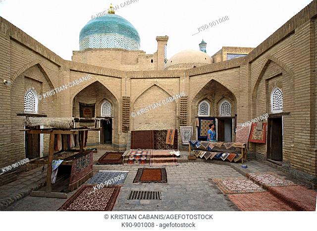 A carpet workshop where handmade silk carpets are displayed inside an old Caravanserai with the Pahlavon Mahmud Mausoleum in the background