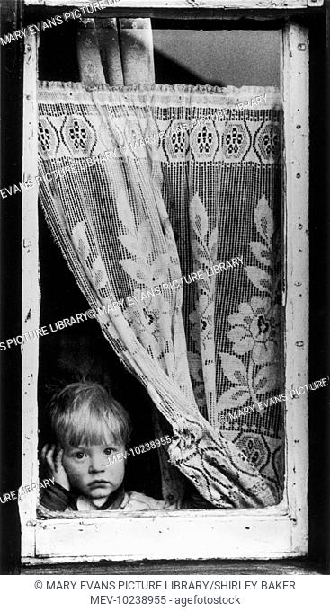 An angelic-faced young boy stares through a gap below the net curtain in a window of a house in Salford, Manchester. Photograph by Shirley Baker