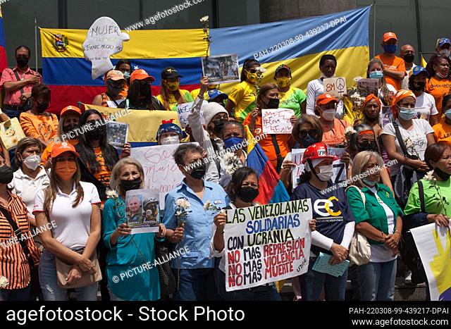 08 March 2022, Venezuela, Caracas: Women with mouth-to-nose coverings hold flowers and placards in front of the flags of Venezuela and Ukraine during a rally...