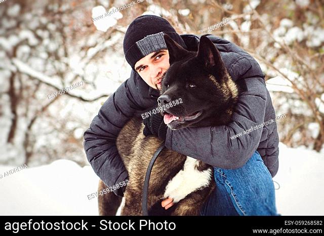 A man in a jacket and a knitted hat walks through a snowy forest with an American Akita dog