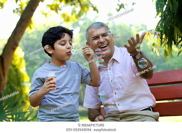Boy blowing bubbles in a park while grandfather looks on