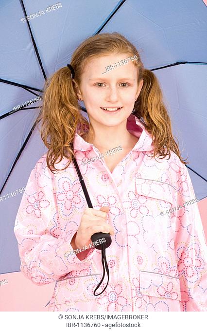 Red-haired girl holding a light blue umbrella and wearing a raincoat in front of a pink backdrop