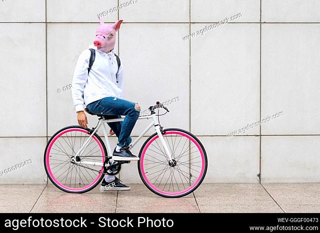 Young man sitting on bicycle with pig mask against wall on footpath