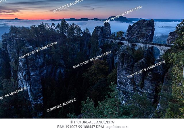 07 October 2019, Saxony, Dresden: In the morning fog lies over the Saxon Switzerland National Park behind the bastion bridge built in 1851 and the...
