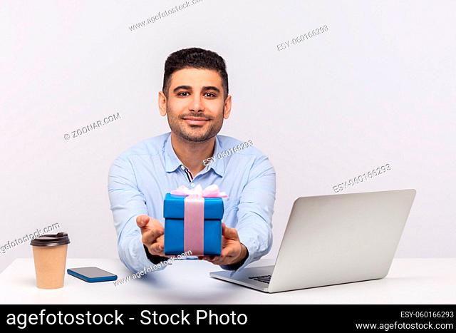 Positive elegant businessman sitting in office workplace, giving gift box to camera and smiling friendly, holding present bonus