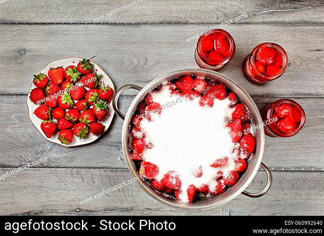 Tabletop view, large steel pot with strawberries covered in crystal sugar, three flasks with fruit in syrup and plate next to it