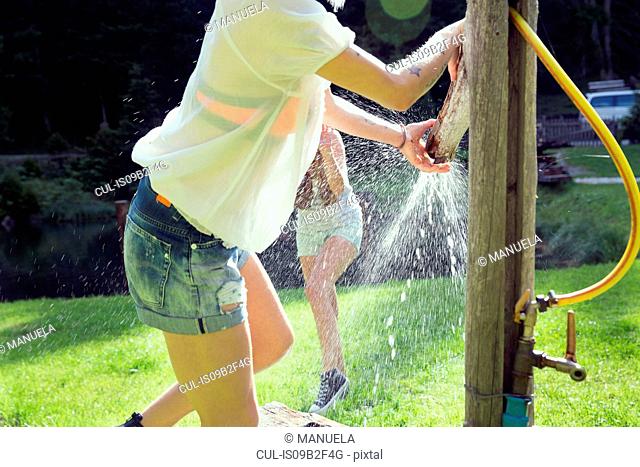 Two adult female friends play fighting sprinkling water hose in garden