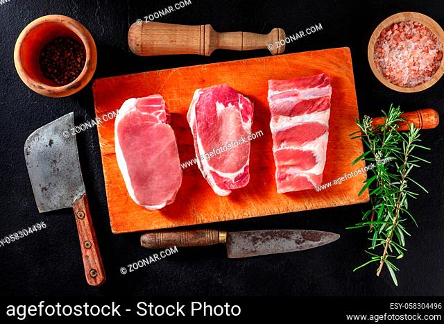 Pork meat, various cuts, overhead flat lay shot on a black background with rosemary, pepper, and knives