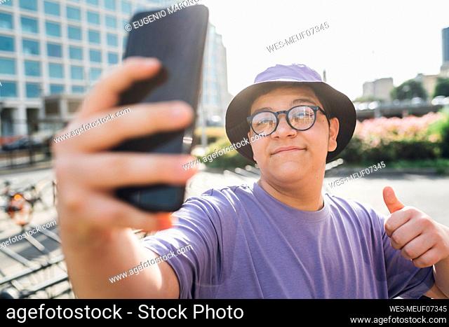 Smiling teenage boy gesturing thumbs up and taking selfie on smart phone