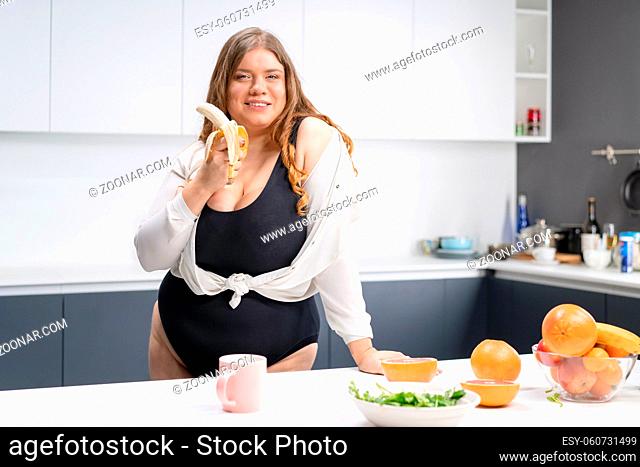 Dieting and nutrition concept. Happy curvy body young woman with long blond hair using blender at modern kitchen, blending fresh fruits for healthy smoothie