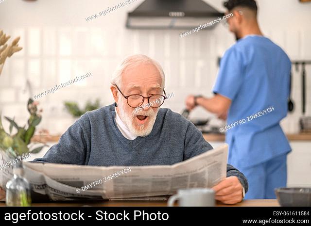 Surprised senior citizen with a newspaper sitting at the kitchen table while his caretaker preparing breakfast