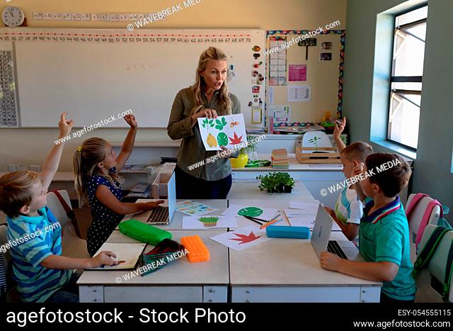 Front view of a Caucasian female teacher with long blonde hair holding a picture of leaves and a diverse group of schoolchildren sitting at desks looking and...