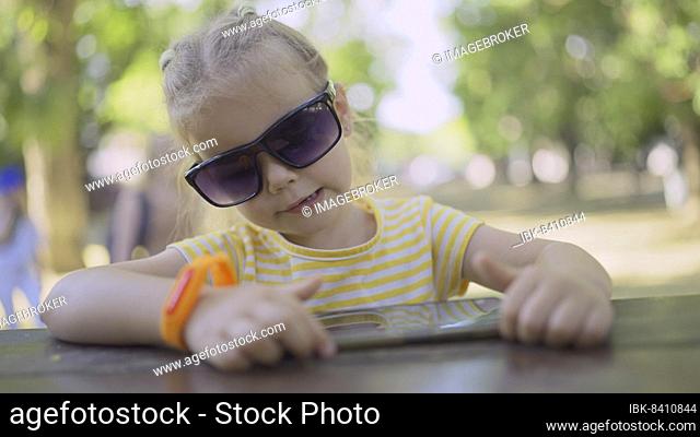 Cute little girl in moms sunglasses listens to music on a mobile phone and sings along. Close-up portrait of child girl sitting in sunglasses on city park and...