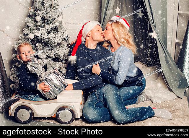 Christmas home portrait of a happy family. Boy holds a gift and sits on a typewriter. Mom and dad in Santa Claus hats kiss each other