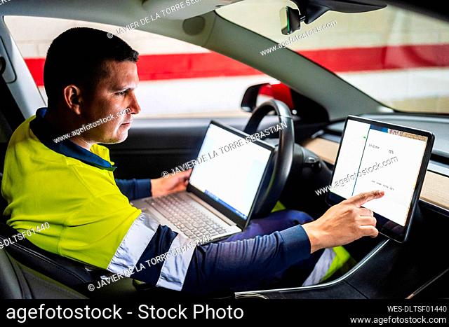 Male programmer with laptop working on digital tablet in electric car