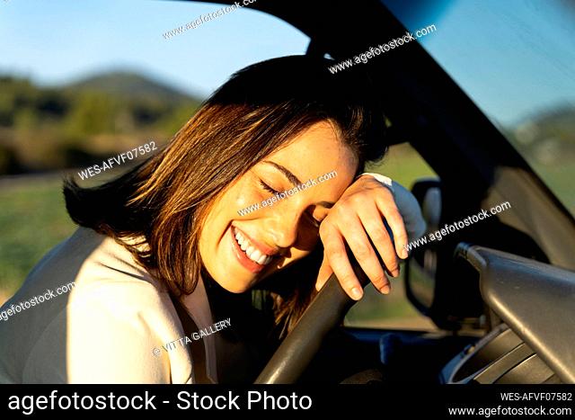 Smiling young woman leaning on steering wheel during sunset