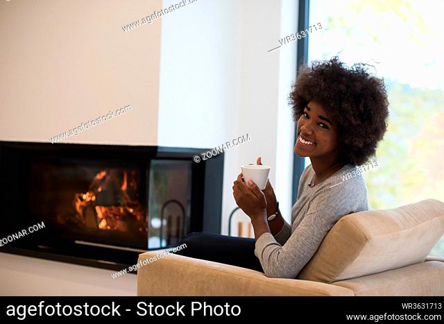 Happy joyful african american woman drinking cup of coffee relaxing at fireplace. Young black girl with hot beverage heating warming up