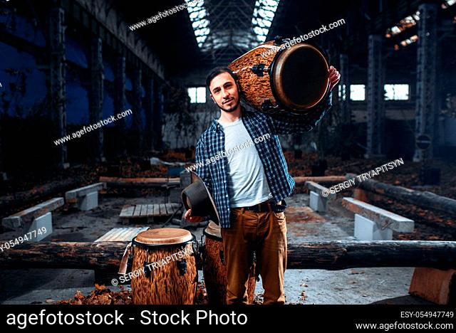 Young male drummer holds wooden drum on the shoulder, factory shop on background. Djembe, musical percussion instrument