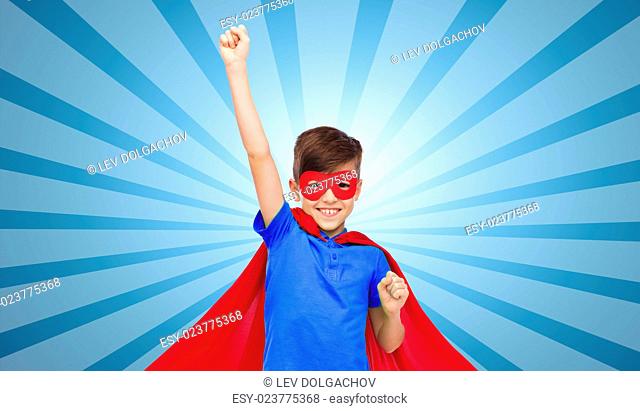 carnival, childhood, power, gesture and people concept - happy boy in red super hero cape and mask showing fists over blue burst rays background