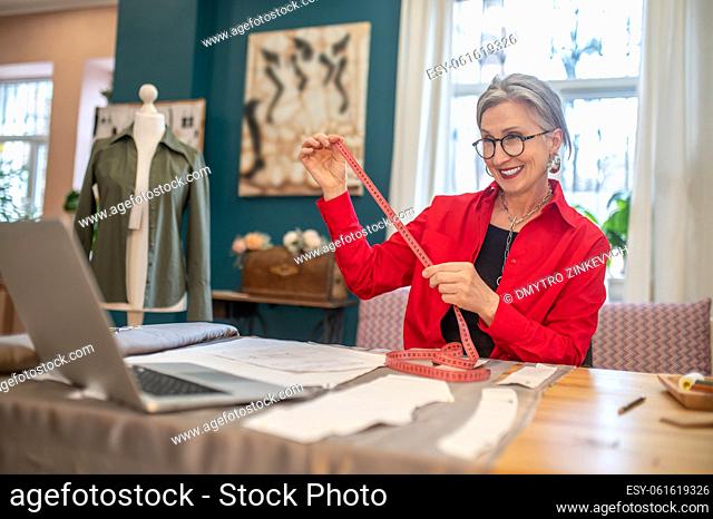 Elegant middle-aged woman in glasses with measure smiling at laptop sitting at table with material and pattern