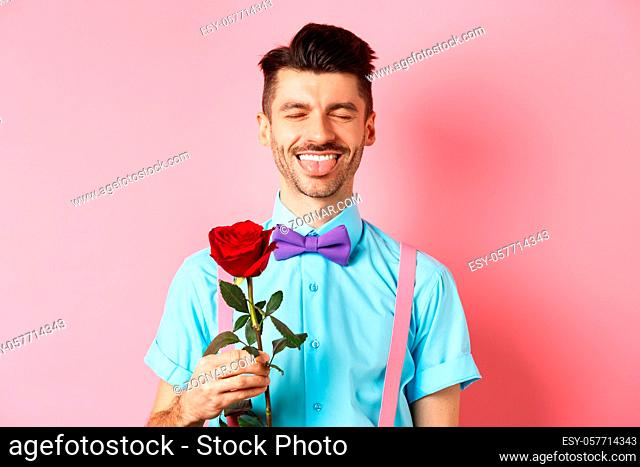 Happy man showing tongue and smiling, holding red rose for girlfriend on Valentines day, enjoying romantic date, pink background
