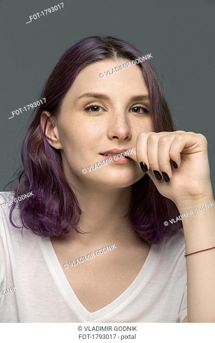 Portrait of a young woman with dyed hair biting nail against gray background