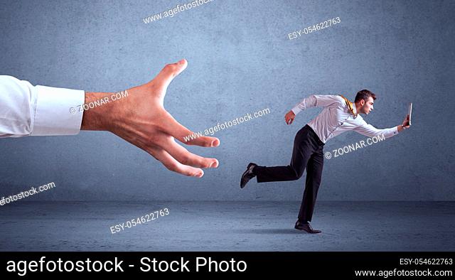 Young miniature businessman running from a big hand with blueish grey background