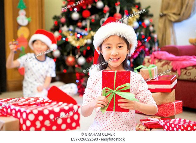 Happy Chinese girl sitting on the floor in her living room at Christmas time. She is surrounded by presents, one of which she is holding with a big smile on her...