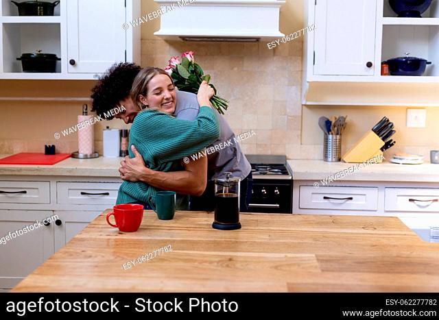 Biracial happy young woman with roses embracing husband while sitting with coffee at table
