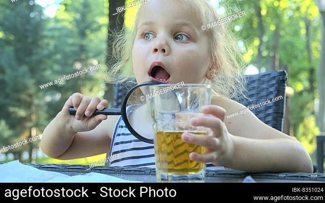 Little girl looks into the lens around. Close-up of blonde girl studying the world around her looking at it through magnifying glass while sitting in street...