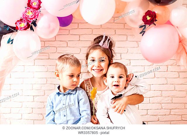 Boys and girl with crowns under birthday balloon and paper flower arch decorations. Childish photozone for celebration