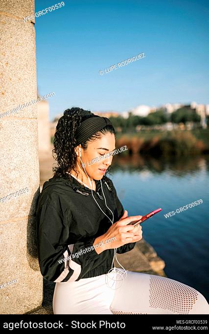 Sportswoman using smart phone and listening music while leaning on column during sunny day