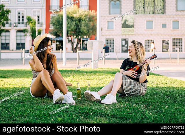Cheerful woman playing ukulele while sitting with female friend on grassy land