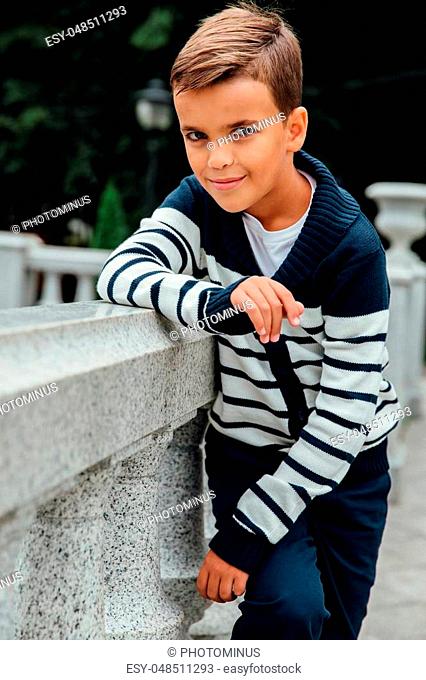 Stylish rich little boy in fashionable clothes. Leaned on Granite-stone handrails. stylish, handsome posing outside in the city park